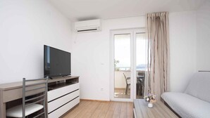A modern, well-lit room with a flat-screen TV, a desk with drawers, a chair, a sofa, and a sliding glass door leading to an outdoor area.