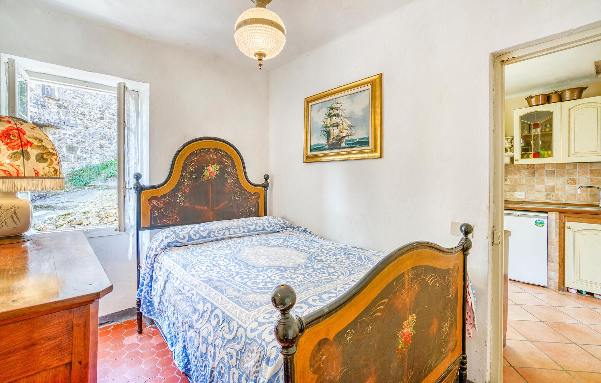 Une chambre avec un lit en bois, une table de chevet, une lampe, une fenêtre donnant sur un mur de pierre et un tableau représentant un navire accroché au mur.