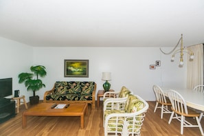 A living room with a wooden coffee table, a sofa, a dining table with chairs, a lamp, and a painting on the wall.