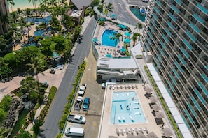 An aerial view of a hotel with multiple swimming pools, a parking area, and a beach area with palm trees.