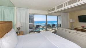 A hotel room with a bed, a nightstand, a lamp, a sofa, a TV, and a balcony with a view of the ocean.