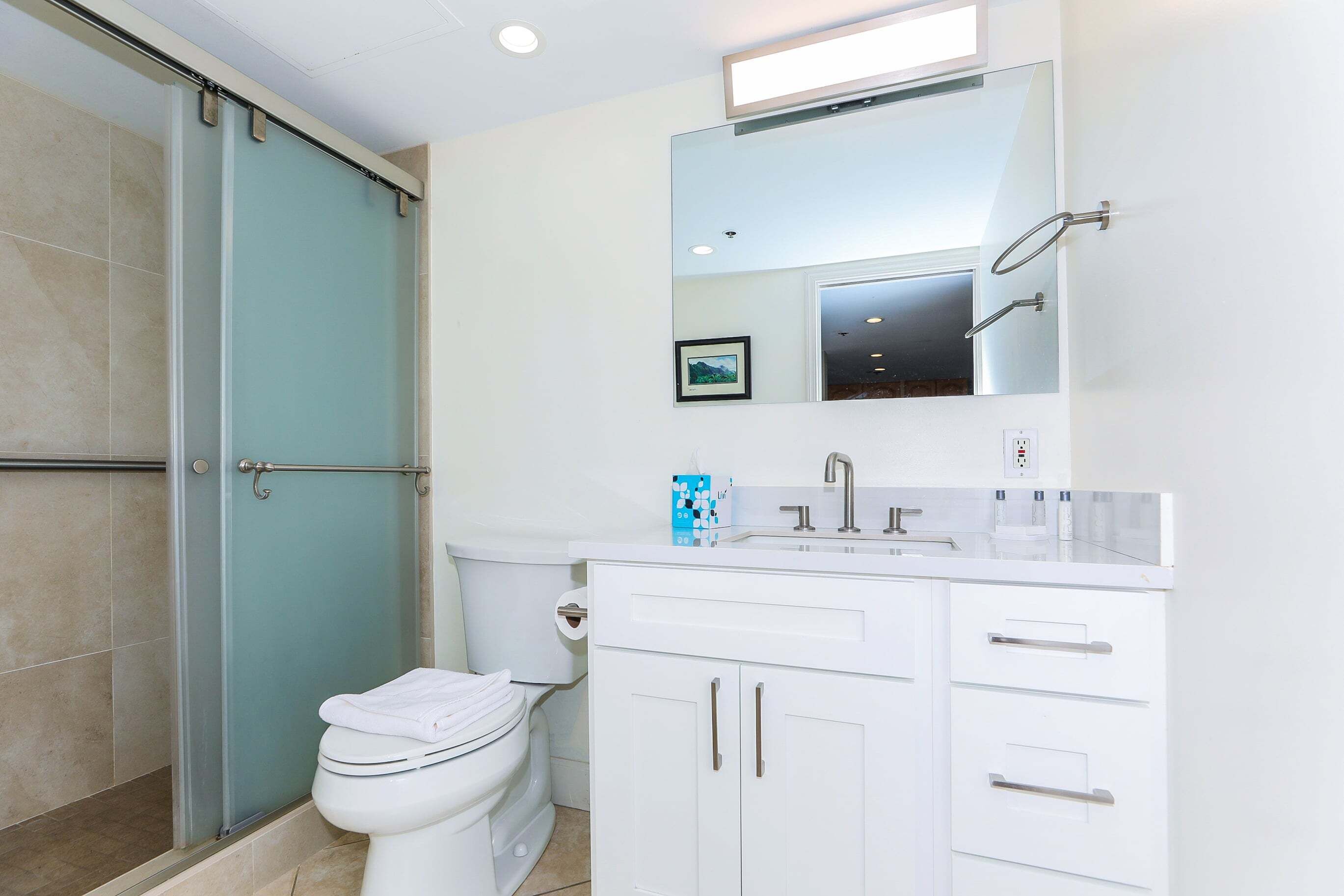 A bathroom with a glass shower enclosure, a toilet, a sink with a mirror, and white cabinets.
