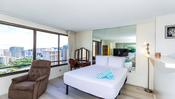A hotel room with a large bed, a reclining chair, a desk, and a city view through large windows. - Spacious Studio, Perfect Location In Wakiki, Located Near The Beach, Shopping (Honolulu)