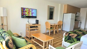 Ett kompakt vardagsrum med en soffa, ett matbord, två stolar, en TV, ett litet köksutrymme med kylskåp och mikrovågsugn samt ett träskåp.