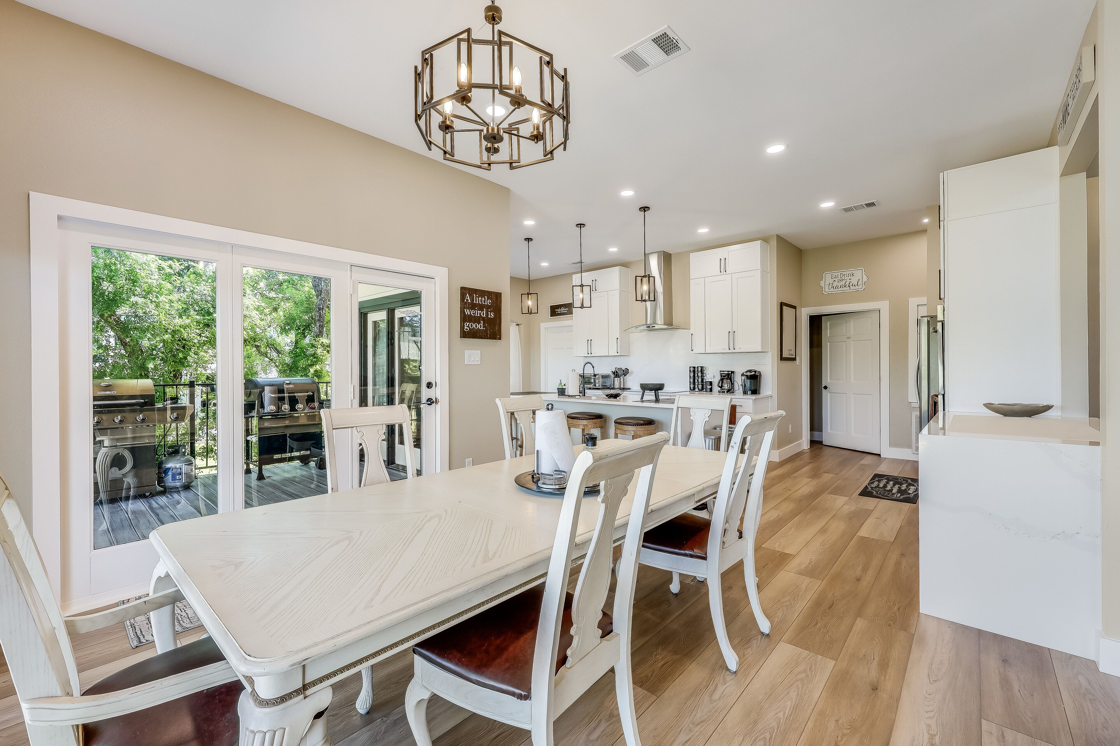 A modern kitchen with white cabinetry, a dining area with a large table and chairs, and a view of the outdoors through glass doors.