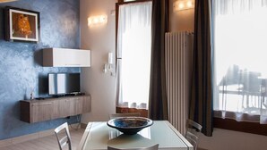 Un comedor moderno con una mesa de vidrio, sillas, un bol, un televisor sobre un mueble y vistas a través de una ventana con cortinas.