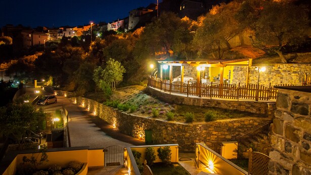 Un edificio de paredes de piedra con una terraza de madera y un área de estar cubierta, rodeado de jardines ajardinados y un sendero sinuoso.
