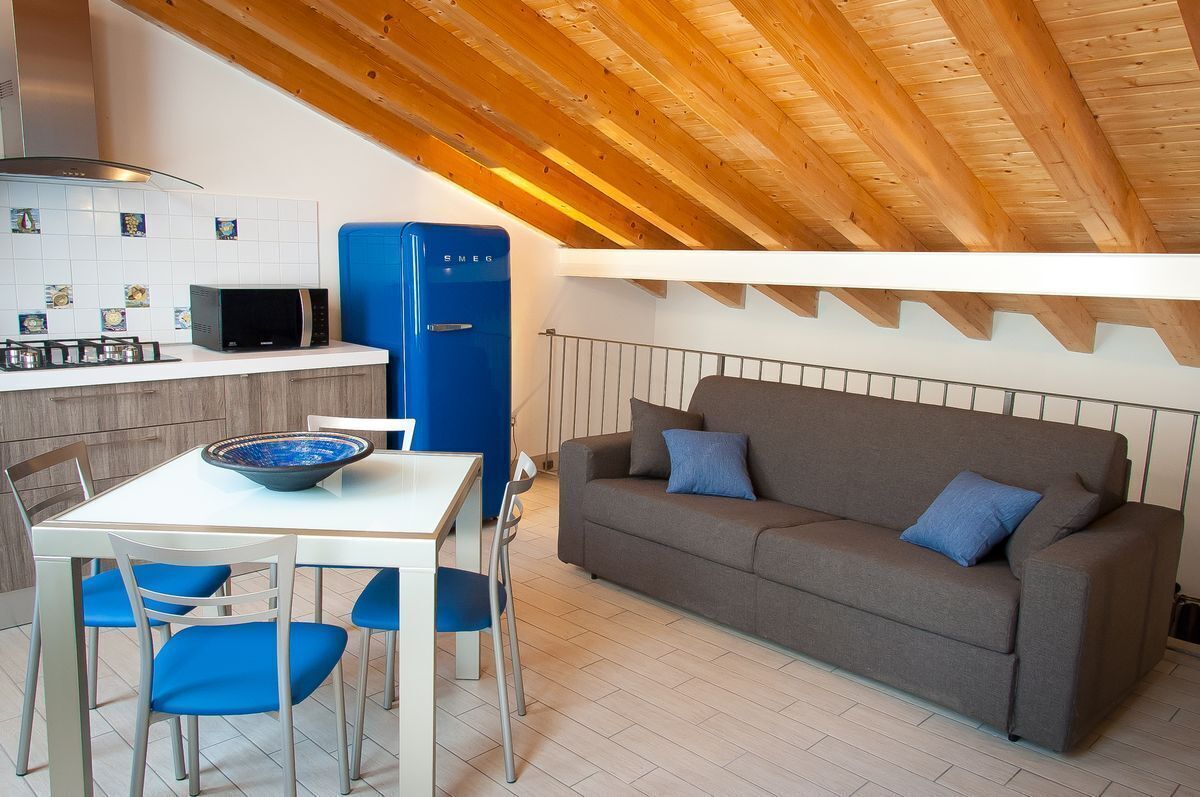 Eine kompakte Küche mit Mikrowelle, Herd und blauem Kühlschrank. Es gibt einen Esstisch mit blauen Stühlen und ein graues Sofa mit blauen Kissen.