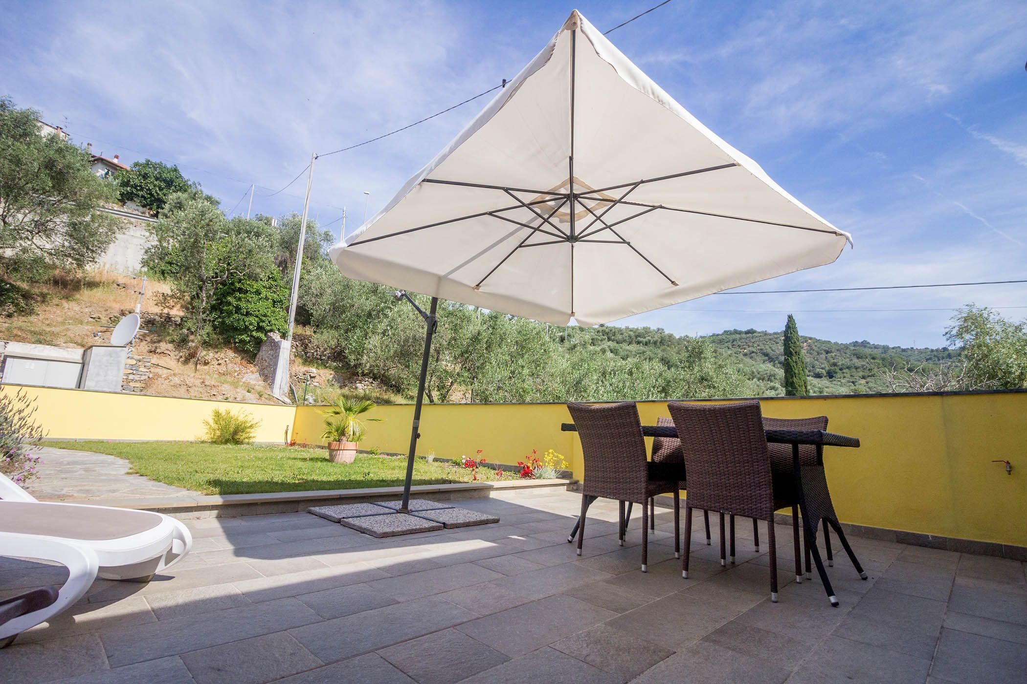 Een terras buiten met een tafel, stoelen, een grote parasol en een gele muur.