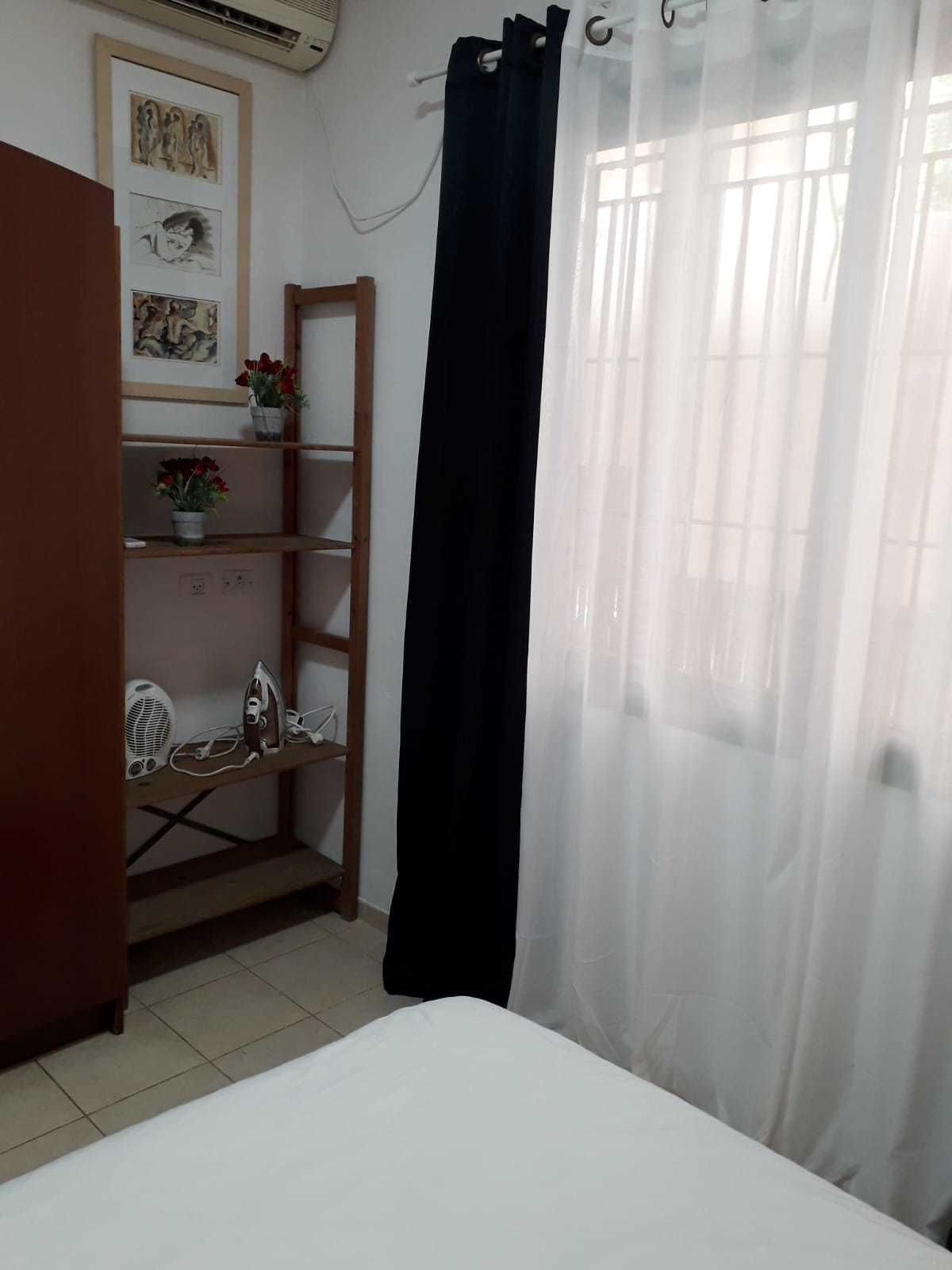 A bedroom with a bed, a wooden shelf with decorative items, a black curtain, and a window with natural light.