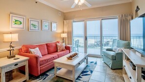 A living room with a red sofa, a white coffee table, a green armchair, and a TV on a wooden stand. There are framed pictures on the wall and a ceiling fan above. - Seawind 402 | Great condo with perfect beach view and walk to dine, shop, more! (Gulf Shores)
