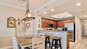 A modern kitchen with wooden cabinets, a central island with stools, and a dining area with a wooden table and chairs. - Seawind 402 | Great condo with perfect beach view and walk to dine, shop, more! (Gulf Shores)
