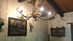 Ein Essbereich mit Holzbalkendecke, einer Kronleuchter mit Geweihen, einem gerahmten Gemälde und einem rustikalen Holztisch.