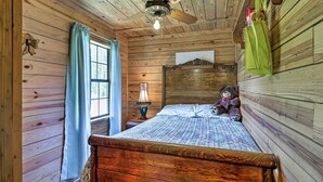 Een slaapkamer in een houten hut met een bed, een plafondventilator, een raam met gordijnen en een teddybeer op het bed.