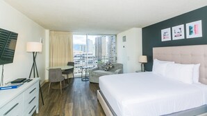 Habitación de hotel con una cama grande, un escritorio con silla, una mesita y vistas a edificios altos por la ventana.
