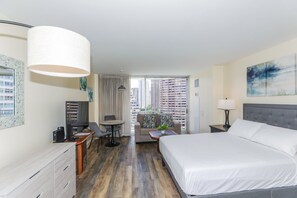 A hotel room with a bed, a desk with a computer, a chair, a small table with a plant, and a view of high-rise buildings through the window.