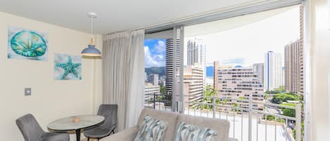 A modern living room with a grey sofa, two grey chairs, a small round table, and a pendant light. There are two wall art pieces, a large starfish and a smaller starfish. A large window with a view of a cityscape and buildings is visible.