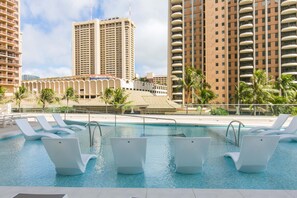 A pool with white lounge chairs, surrounded by tall buildings and palm trees.