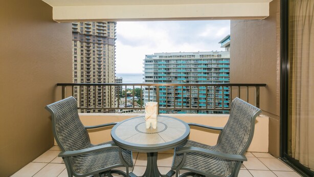 A balcony with a round table and two chairs, overlooking a cityscape with high-rise buildings. - Great Location In Wakiki, Walking Distance To Beach, Shopping, Dining! (Honolulu)