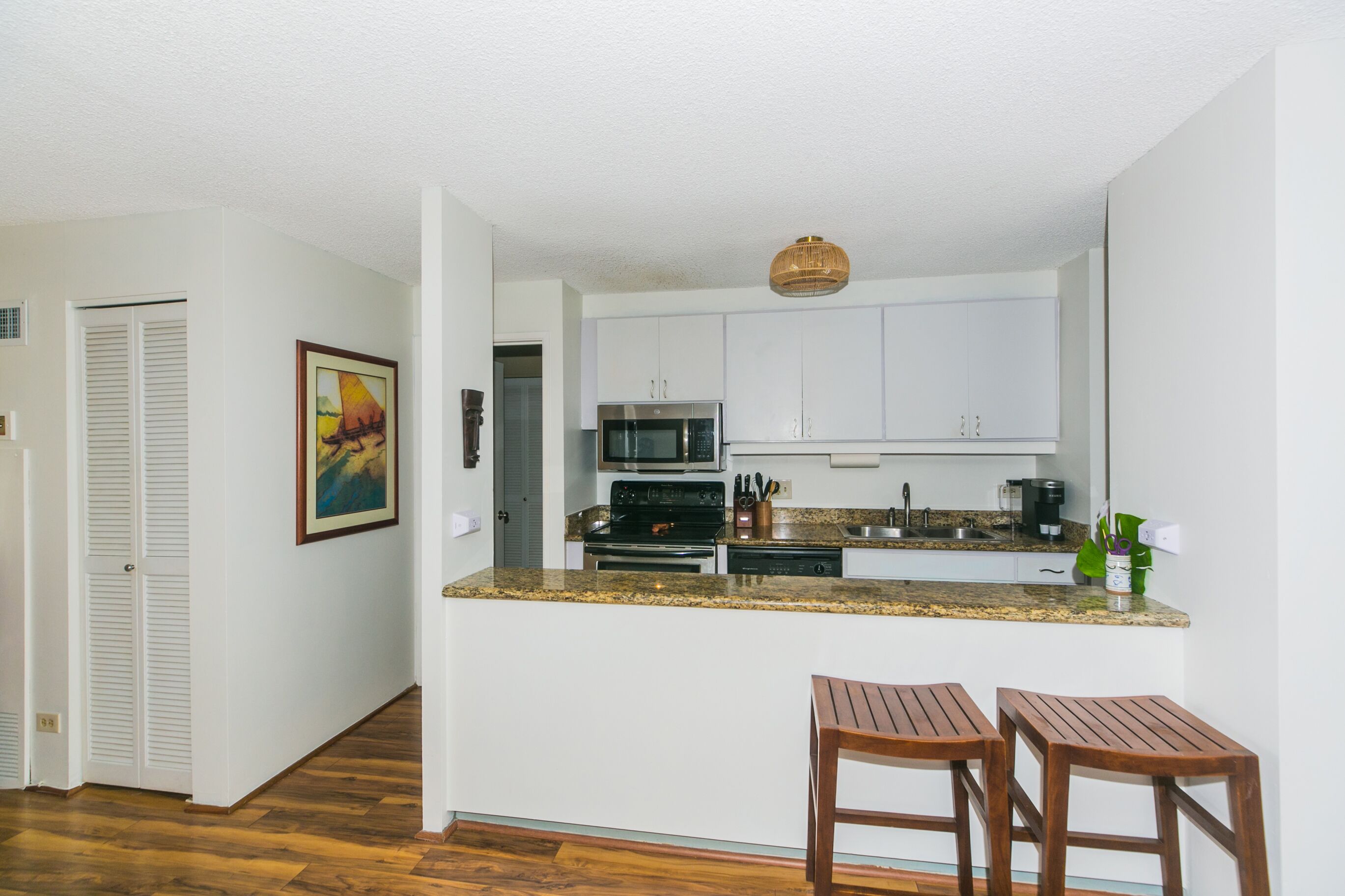 A compact kitchen with white cabinets, a granite countertop, a microwave, a stove, and a sink. There are two wooden stools at the counter.