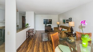 A modern living room with a wooden floor, a glass dining table, a sofa, and a TV mounted on the wall. - Great Location In Wakiki, Walking Distance To Beach, Shopping, Dining! (Honolulu)