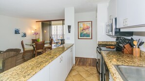 A kitchen with granite countertops, white cabinets, and a dining area with wicker chairs and a table. - Great Location In Wakiki, Walking Distance To Beach, Shopping, Dining! (Honolulu)