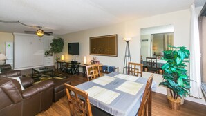 Ein Wohnzimmer mit einer Couch, einem Couchtisch und einem Esstisch, gedeckt mit Unterlagen. Es gibt einen Deckenventilator, einen Fernseher und eine Pflanze im Raum.
