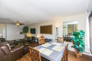 Un salon comprenant un canapé, une table basse et une table à manger avec des sets de table. On y trouve également un ventilateur de plafond, un téléviseur et une plante.