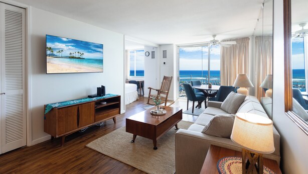 A modern hotel room with a large TV, a sofa, a dining area, and a balcony with a view of the ocean.