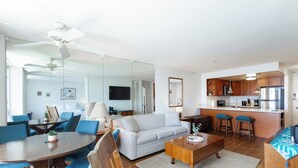 A modern living room with a sofa, coffee table, and blue chairs. A kitchen with wooden cabinets and stainless steel appliances is visible in the background.