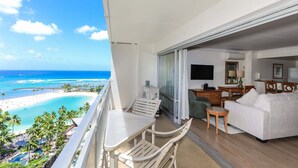 Een balkon met uitzicht op een strand en de oceaan, een eethoek met tafel en stoelen, en een woonkamer met een bank en een televisie.