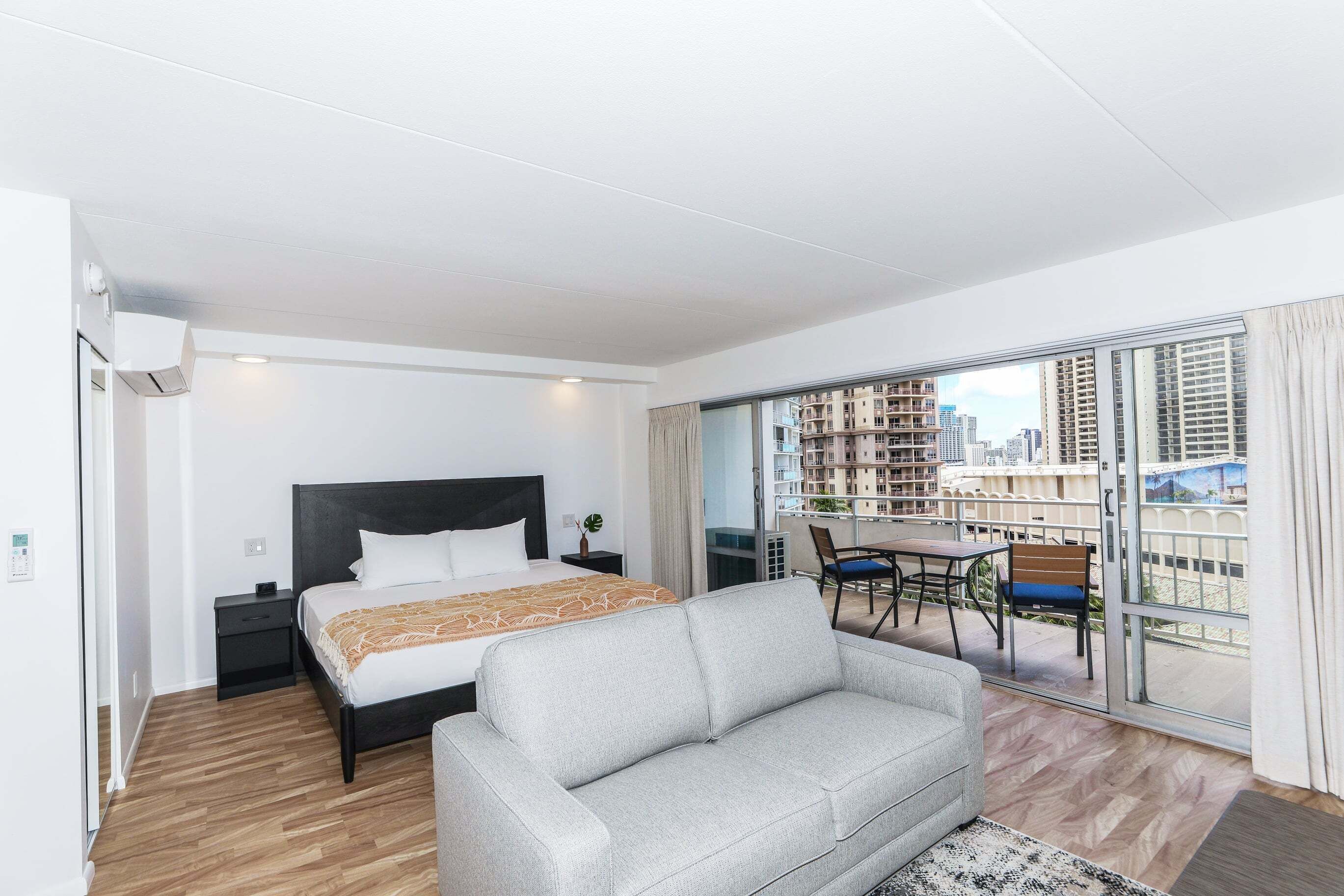 A modern hotel room with a bed, a sofa, a balcony with a table and chairs, and a view of the cityscape.