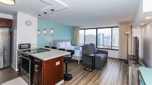 A modern hotel room with a kitchenette, a bed, a seating area, and a large window offering a city view.