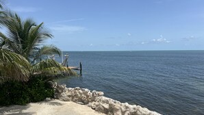 A coastal view with a concrete walkway, palm trees, and a distant sailboat on the water. - Caribbean Sunsets: 3BR Home Gulf Views Pool & Dock (Marathon)