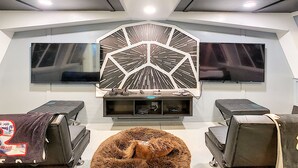 Ein modernes Wohnzimmer mit zwei großen Flachbildfernsehern an der Wand, einem zentralen Wandkunstwerk und einer gemütlichen Sitzecke mit einem runden, flauschigen Pouf.