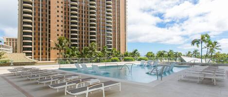A hotel pool area with lounge chairs, a swimming pool, and a tall building in the background.