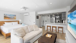 Een moderne hotelkamer met een bed, bank, bijzettafeltje, keukenhoek en een groot televisiescherm.