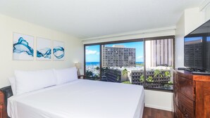 A hotel room with a large bed, a TV, a wooden dresser, and a view of a cityscape and beach. - Relaxing, Spacious, Great Location In Waikiki! (Honolulu)