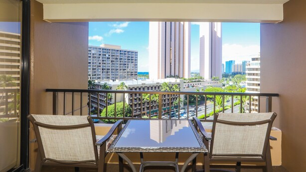 A balcony with two wicker chairs and a glass table, overlooking a cityscape with tall buildings and palm trees. - Relaxing, Spacious, Great Location In Waikiki! (Honolulu)