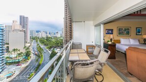 Un balcón con mesa y sillas, una cama y vistas a un paisaje urbano con edificios altos y palmeras.