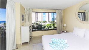 Une chambre d’hôtel avec un grand lit, un bureau, une chaise, une lampe, un miroir rond et une fenêtre donnant sur des immeubles et des palmiers.