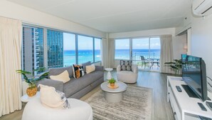 Una sala de estar moderna con un amplio ventanal con vistas al océano, un sofá gris, un sillón blanco y un televisor de pantalla plana sobre un mueble blanco.