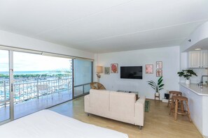 A modern living room with a balcony overlooking a marina, a flat-screen TV, and a kitchen area with bar stools. - Highly desirable Deluxe One-bedroom Yacht Harbor View - Reserve Today, Will not last Long!! (Honolulu)