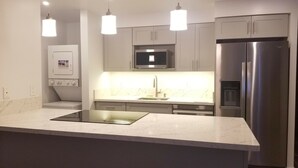 Cozinha moderna com geladeira de aço inoxidável, micro-ondas e forno embutido. Há luminárias pendentes sobre a bancada, e os armários são de cor clara.