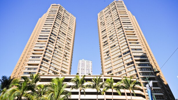 Dois edifícios altos com varandas, palmeiras ao primeiro plano e um céu azul límpido.
