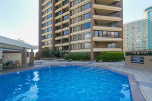 Une piscine dont le bord est recouvert de carreaux, entourée d’un bâtiment doté de nombreux balcons et d’un panneau indiquant qu’il n’y a pas de sauveteur en service.