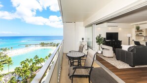 Een balkon met uitzicht op een strand en de oceaan, een eettafel met stoelen, een bank en een televisie in de woonkamer.