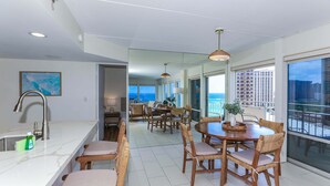 A modern kitchen with a large island, dining area with wooden chairs, and large windows offering a view of the city. - Ilikai Hotel, Amazing Ocean views! Most desirable location in Ilikai! (Honolulu)
