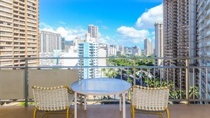 A balcony with a round table and two wicker chairs, overlooking a cityscape with high-rise buildings and a clear sky. - Ilikai Hotel - No Resort Fees!  City-views, condo on the 14th Floor,  Prime Waikiki location! (Honolulu)