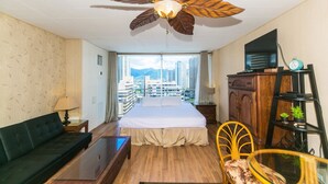 A hotel room with a bed, a sofa, a TV, a ceiling fan, and a view of the city.
